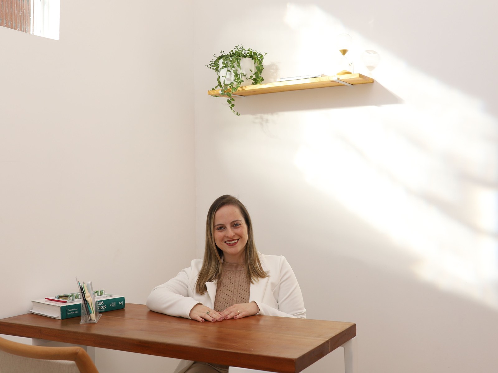Dra. Israella em blazer branco no consultório, apoiada em mesa de madeira, com prateleira iluminada pela luz do sol